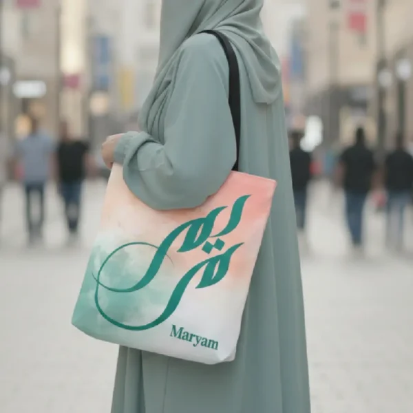 Maryam name in BinMahmood original Arabic fusion calligraphy in teal on a polyester tote bag, carried by a modest woman in abaya and hijab — mouse-crafted by BinMahmood