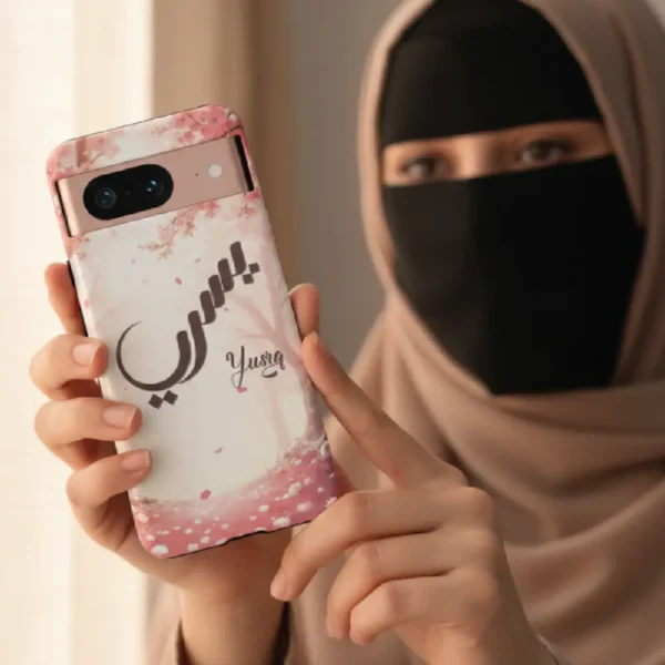 A modest lifestyle view of a woman with a niqab and hijab holding a pink cherry blossom phone case featuring the Yusra bespoke Arabic calligraphy name art by BinMahmood.