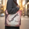 A modest female with a hijab carrying the Yusra bespoke Arabic name canvas tote bag over her shoulder to show real world size by BinMahmood.