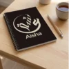 Product photo shoot of Aisha Arabic calligraphy notebook, lay flat on a white pine table with a pen and a coffee mug in the surrounding - binmahmood