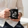 Product photoshoot of Arqam personalized Arabic calligraphy black coffee mug, held by a model in hands wearing an off white linen shirt and black cotton chinos - binmahmood