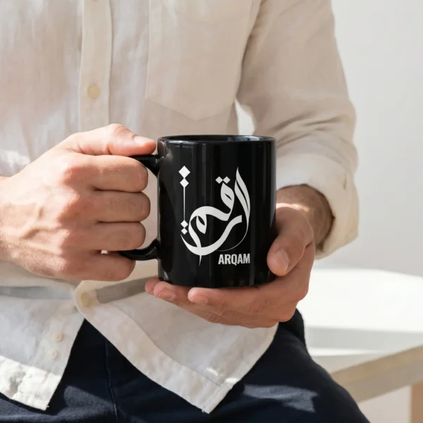 Product photoshoot of Arqam personalized Arabic calligraphy black coffee mug, held by a model in hands wearing an off white linen shirt and black cotton chinos - binmahmood