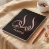Product photoshoot of Asma name Arabic calligraphy notebook lay flat on a beige linen over wooden table with a pen and dried flower - binmahmood