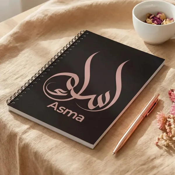 Product photoshoot of Asma name Arabic calligraphy notebook lay flat on a beige linen over wooden table with a pen and dried flower - binmahmood