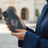 Woman with henna in hands holding a dark grey tough mobile phone case featuring white and red 'Bedouin Love' Arabic calligraphy in a geometric diamond design, with a soft-focus architectural background
