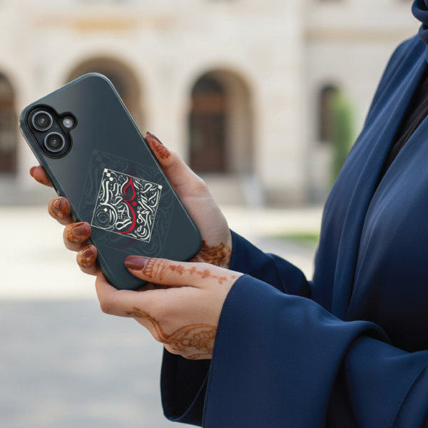 Woman with henna in hands holding a dark grey tough mobile phone case featuring white and red 'Bedouin Love' Arabic calligraphy in a geometric diamond design, with a soft-focus architectural background