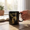 A glossy black ceramic mug featuring gold Fatimah Arabic calligraphy sitting on a dark oak wooden desk in a modern, ambient-lit room.