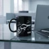 The 11oz mug sits on a glass-top office desk next to a closed MacBook and a slim notebook with a pen resting across it.