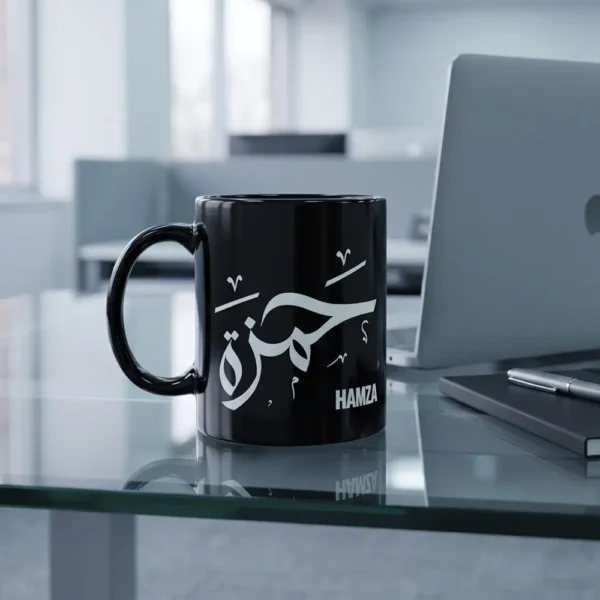 The 11oz mug sits on a glass-top office desk next to a closed MacBook and a slim notebook with a pen resting across it.