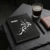 Product photoshoot of Hashim personalized Arabic name calligraphy notebook, lay flat on a placed on a dark slate surface with a minimal industrial feel - binmahmood