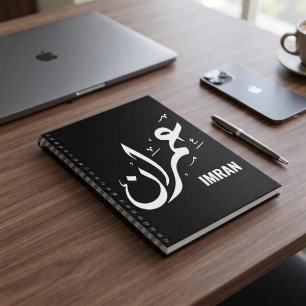 Product photoshoot of Imran Arabic Diwani calligraphy name art spiral notebook, lay flat on a dark wood table, a closed apple laptop and an i phone along with a pen can be seen in the blur background - binmahmood