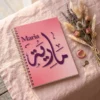 Product photography of Maria Arabic calligraphy name art spiral notebook, lay flat on a soft blush-pink linen surface a small bouquet of dried flowers nearby - binmahmood