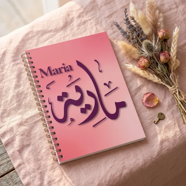 Product photography of Maria Arabic calligraphy name art spiral notebook, lay flat on a soft blush-pink linen surface a small bouquet of dried flowers nearby - binmahmood