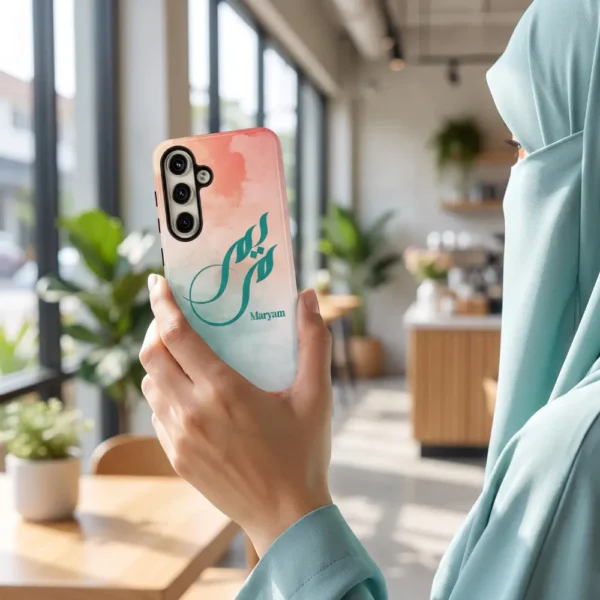 Lifestyle photo of a personalized Maryam Arabic fusion calligraphy phone case held in one hand by a woman wearing a light teal hijab, photographed from behind in a bright modern café.
