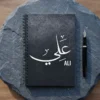 Product photoshoot of Ali personalized Arabic name Thuluth calligraphy spial notebook place on a slate colour hard surface with a pen - binmahmood