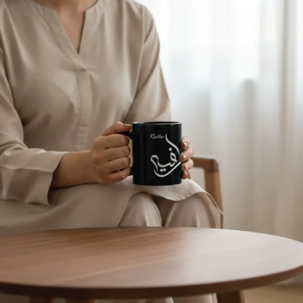 A model in a cream-colored outfit holding a glossy black ceramic mug featuring the name Rafia in white Al Wissam Arabic calligraphy. The design is precision mouse-crafted and displayed in a minimalist lifestyle setting.