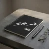Product photo shoot of Saad personalized Arabic calligraphy notebook, lay flat on a charcoal industrial table top with a pen and a glasses - binmahmood