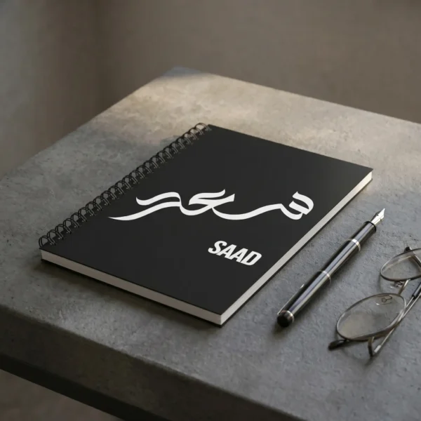Product photo shoot of Saad personalized Arabic calligraphy notebook, lay flat on a charcoal industrial table top with a pen and a glasses - binmahmood
