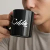 Black ceramic mug with “Shayan” Arabic calligraphy held by male model sipping coffee, face cropped and blurred, minimalist lifestyle setting