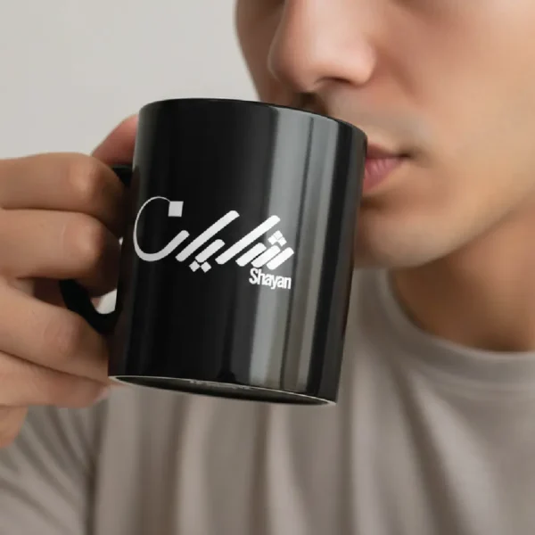 Black ceramic mug with “Shayan” Arabic calligraphy held by male model sipping coffee, face cropped and blurred, minimalist lifestyle setting