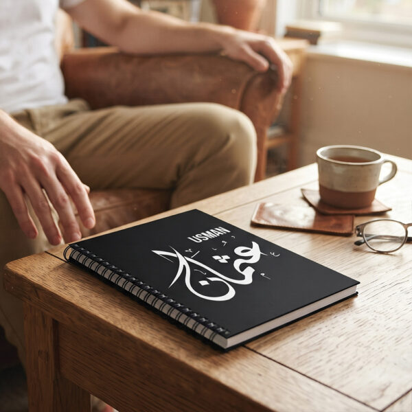 Product photography of Usman Arabic Diwani calligraphy name art spiral notebook, lay flat on a metallic office table - binmahmood