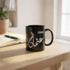 Product View of Usman Arabic name mug in black ceramic with white Diwani calligraphy, on a white oak table with a notebook and an open fountain pen crafted by binmahmood