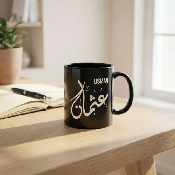 Product View of Usman Arabic name mug in black ceramic with white Diwani calligraphy, on a white oak table with a notebook and an open fountain pen crafted by binmahmood