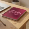 Product photography of Zahra name Arabic Fusin Calligraphy Notebook, lay flat on a pine office table with a pen and a laptop in the background
