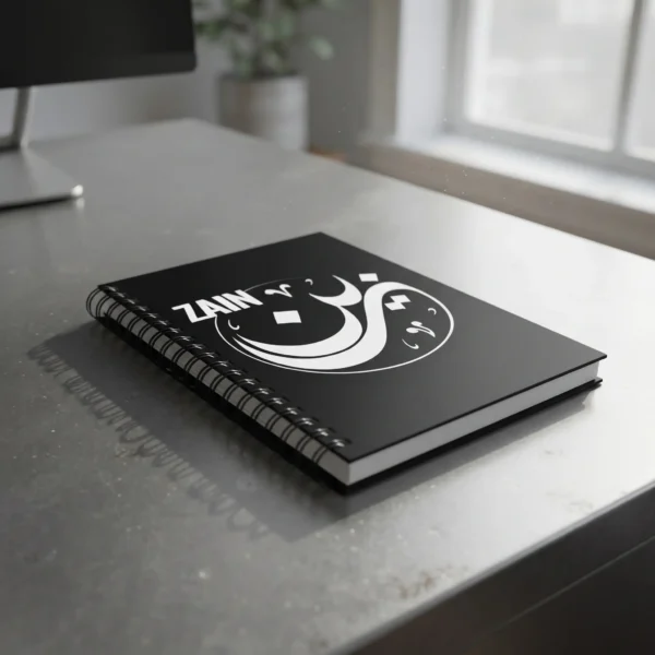Product photography of Zain Arabic Diwani calligraphy name art spiral notebook, lay flat on a metallic office table - binmahmood