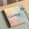 Product photoshoot of Zainab custom Arabic Al Wissam calligraphy notebook, lay flat on a soft sage-green woven placemat with a dried flower - binmahmood