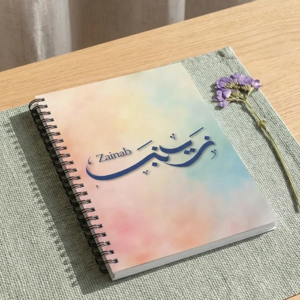 Product photoshoot of Zainab custom Arabic Al Wissam calligraphy notebook, lay flat on a soft sage-green woven placemat with a dried flower - binmahmood